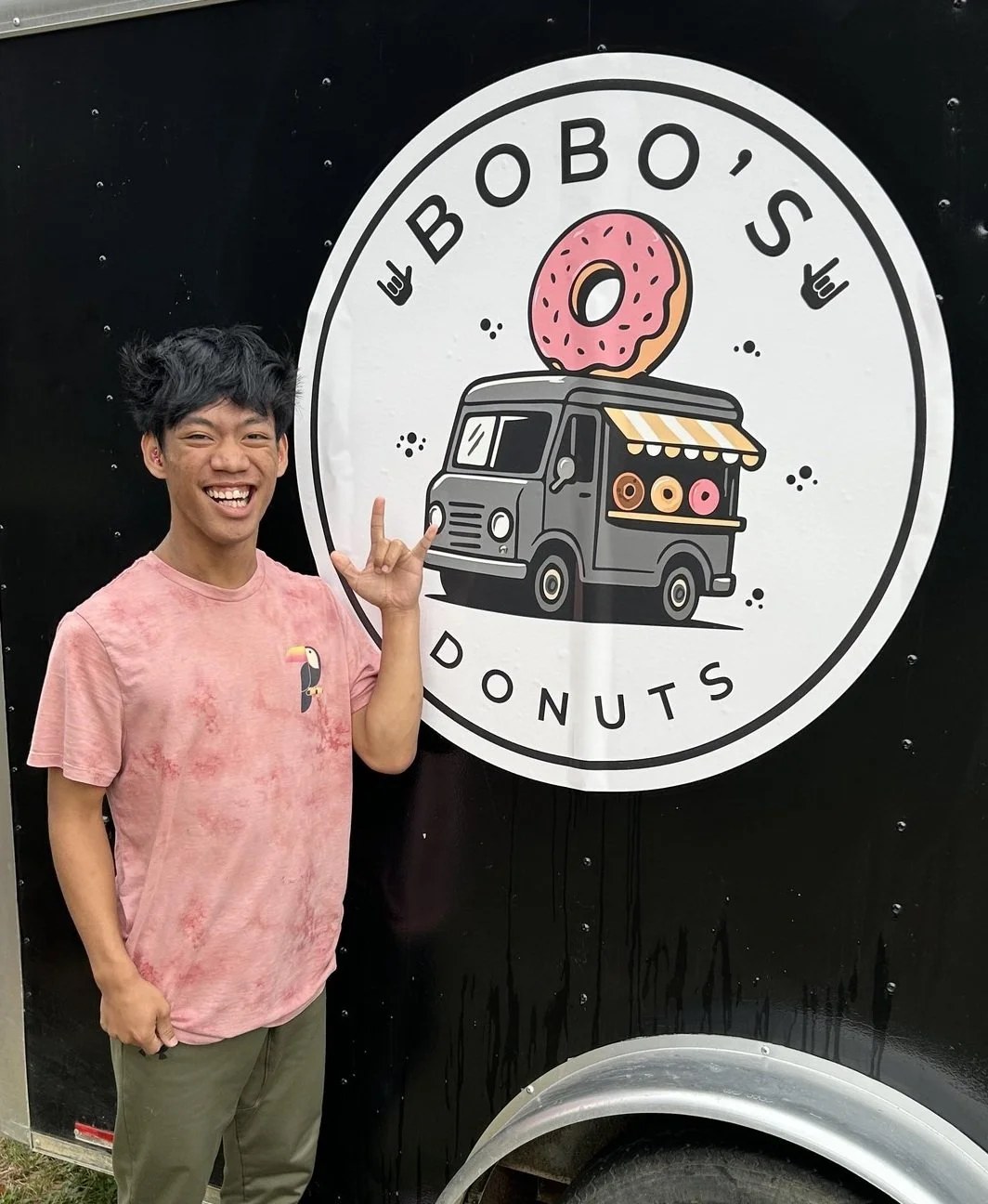 A young man in a pink tie-dye T-shirt smiling and making a hand gesture, standing in front of a black food truck with a sign that says "Bobo's Donuts" and features a cartoon donut on a van.